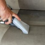 Hands using a vacuum nozzle to deeply clean a fabric car seat, removing dirt and stains.